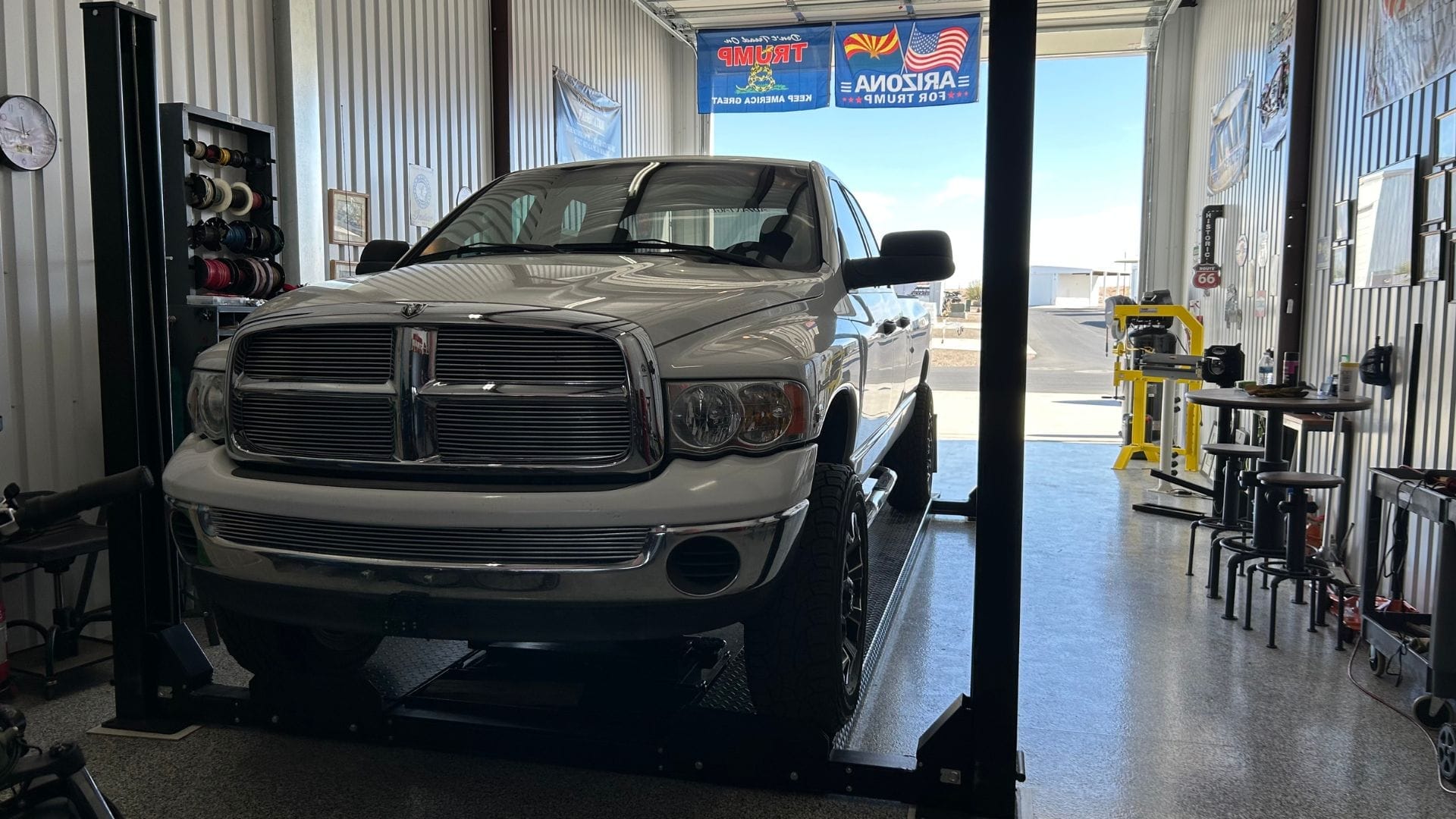 Gallery A white Dodge Ram truck parked on a PP9 car lift in a well-organized garage with tools and banners visible in the background.