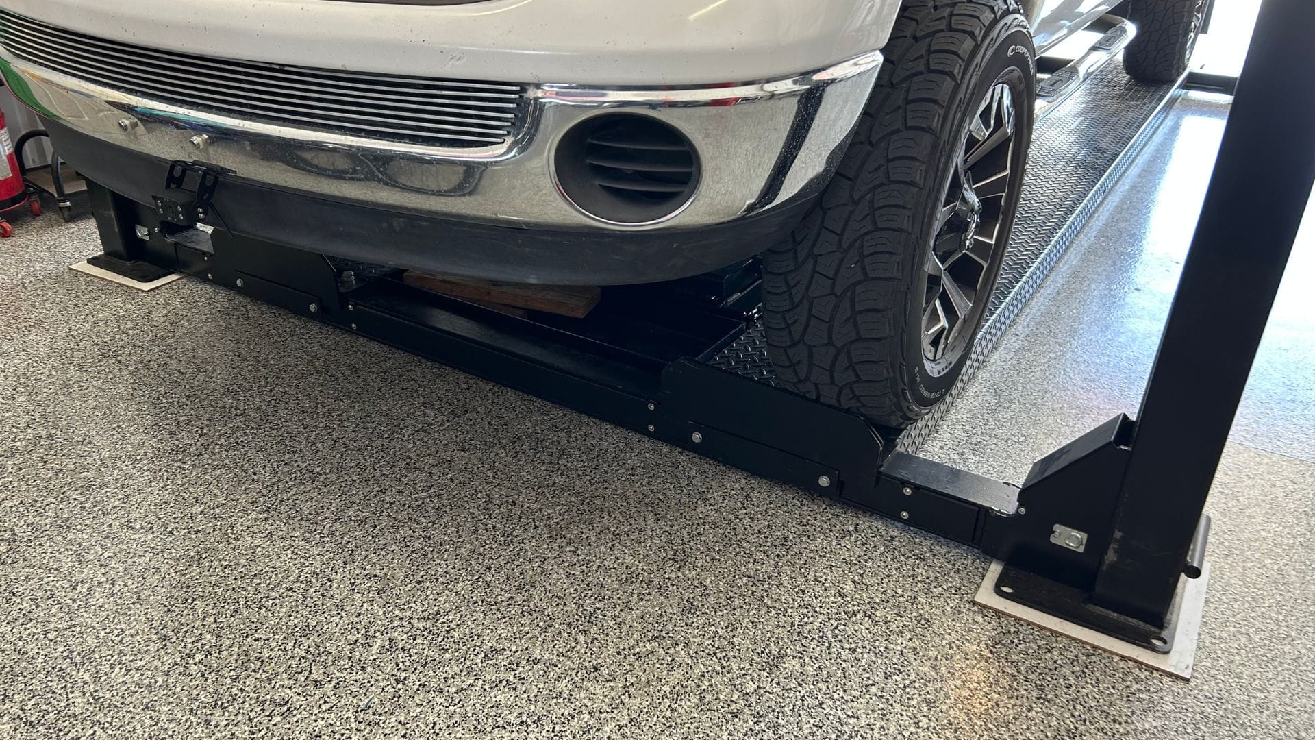 Gallery Close-up of a white Dodge Ram truck's front bumper and wheels positioned on a PP9P car lift with a textured garage floor underneath.