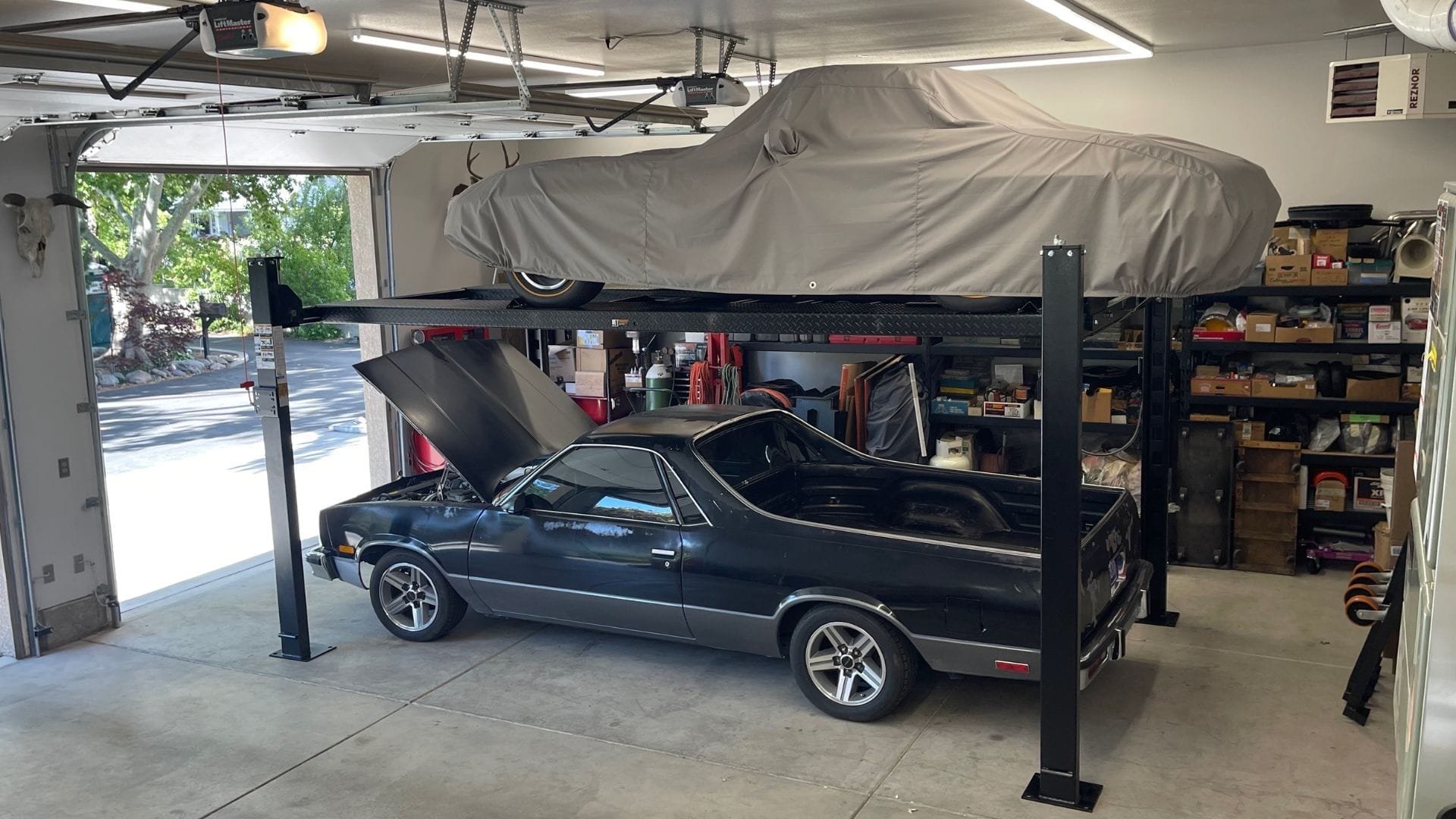 Gallery classic black car with its hood open, parked underneath a covered vintage car on a two-post PP8S car lift in a residential garage.
